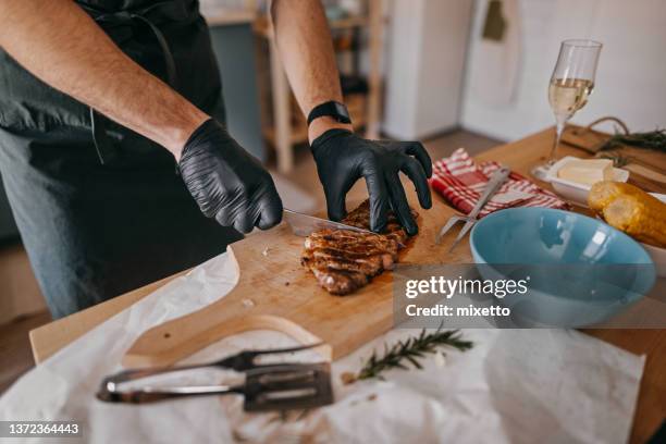 hands of man cutting grilled steak on board in kitchen - zwarte handschoen stockfoto's en -beelden