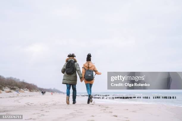 couple of man and woman in warm clothes, active running down the sandy seaside. gray sky, groyne. rearview - romantic couple walking winter beach stock pictures, royalty-free photos & images