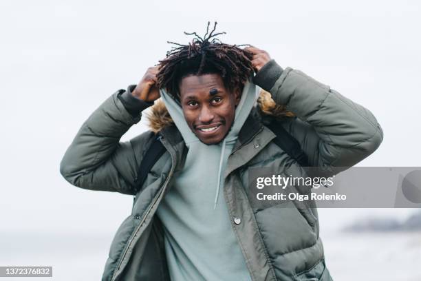 portrait of carefree man alone spending time at windy cold beach. sea tide and gray sky on background - huva bildbanksfoton och bilder