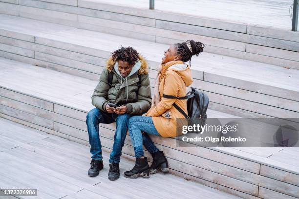 laughing smiling joyful students, woman and man checking smartphone video and photos, sitting on wooden bench. - friends laughing at iphone video stock pictures, royalty-free photos & images