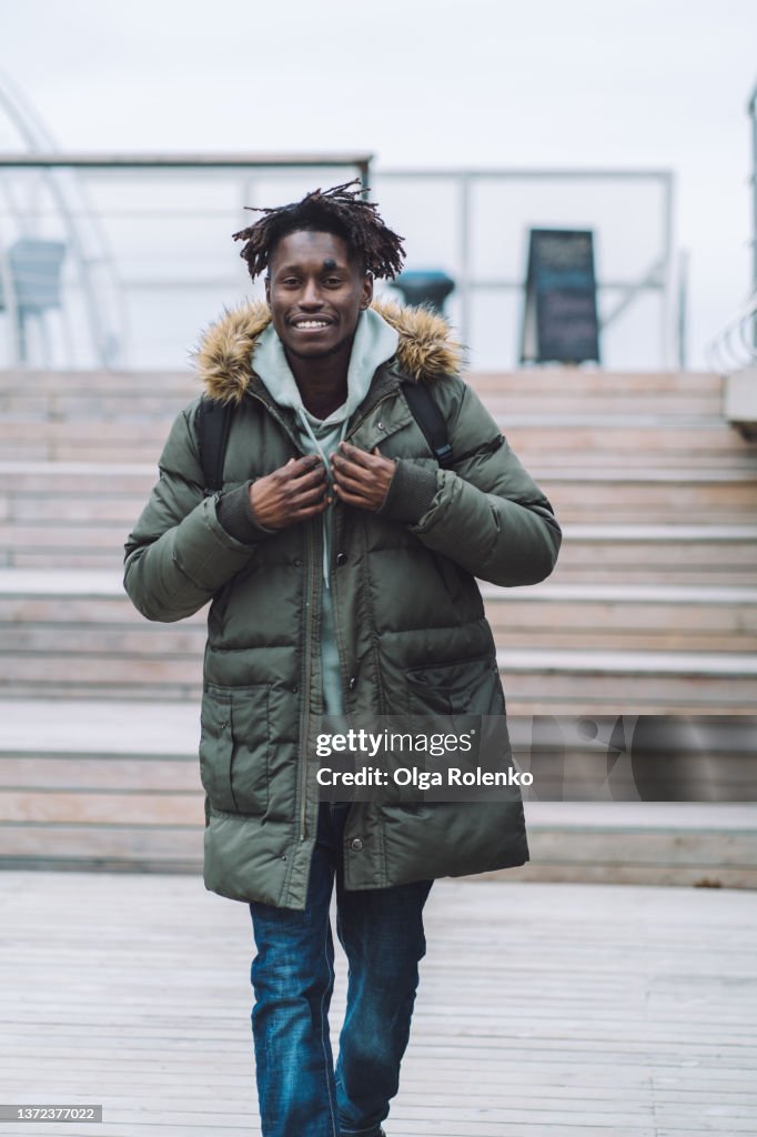 Happy smiling fashionable man in warm clothes and backpack walking on street near wooden bench.