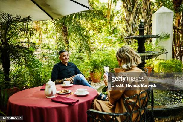 Medium wide shot of woman taking photo of boyfriend with smart phone while have tea in garden of luxury tropical hotel