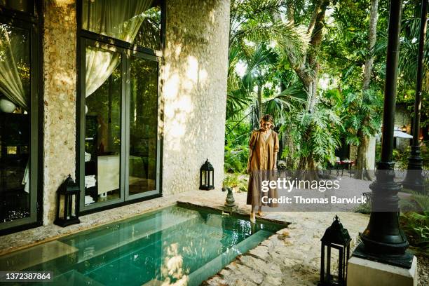 wide shot of smiling woman standing pool and putting foot in water at luxury tropical spa - travel accommodation stock-fotos und bilder