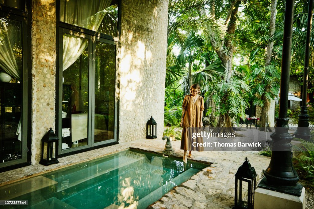 Wide shot of smiling woman standing pool and putting foot in water at luxury tropical spa