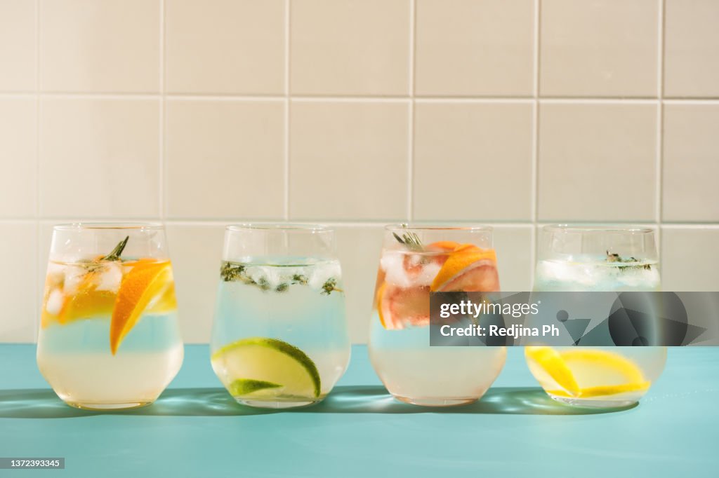 Summer refreshing sparkling drink with ice, fresh fruits and herbs: rosemary, thyme, orange, lime, lemon, grapefruit in transparent glasses on a blue background.