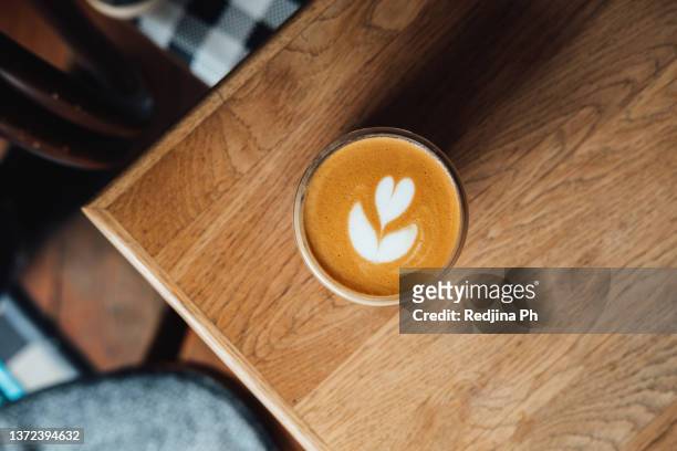 glass cup with coffee and ice latte in the form of  flower and  heart stands on the corner of wooden table. flat white coffee drink. - table top view stock pictures, royalty-free photos & images