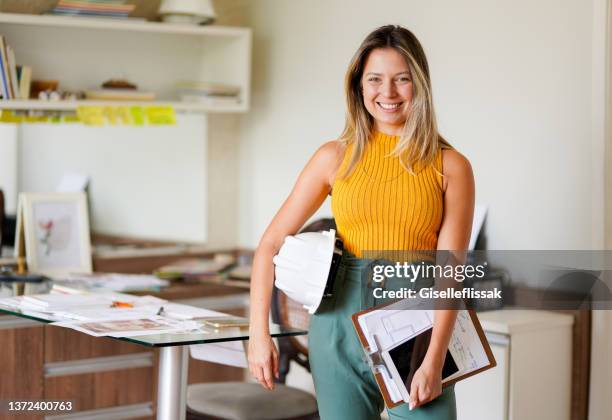 sorridente engenheira feminina em pé com um chapéu duro e prancheta em seu escritório - designer de interiores - fotografias e filmes do acervo