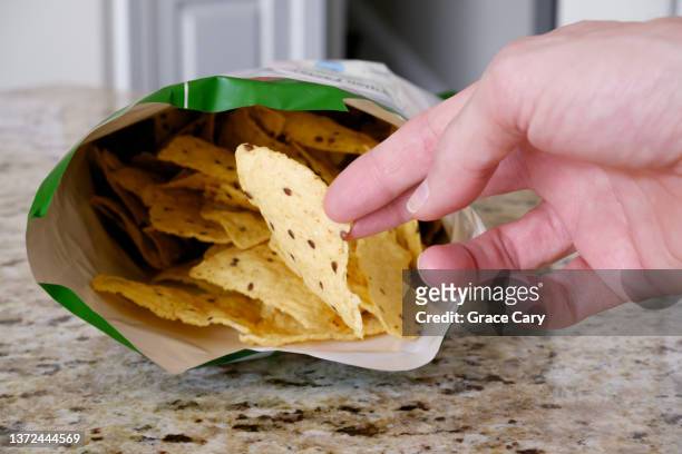 man grabs a tortilla chip from open bag on kitchen counter - bag of chips fotografías e imágenes de stock