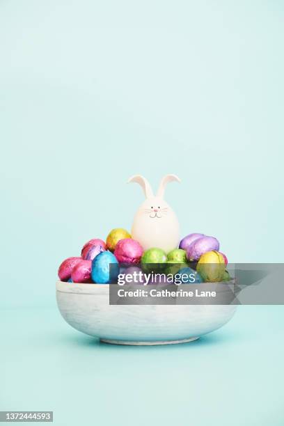 easter background with whitewashed wooden bowl filled with foil covered chocolate easter eggsand cute easter bunny - candy dish stock pictures, royalty-free photos & images