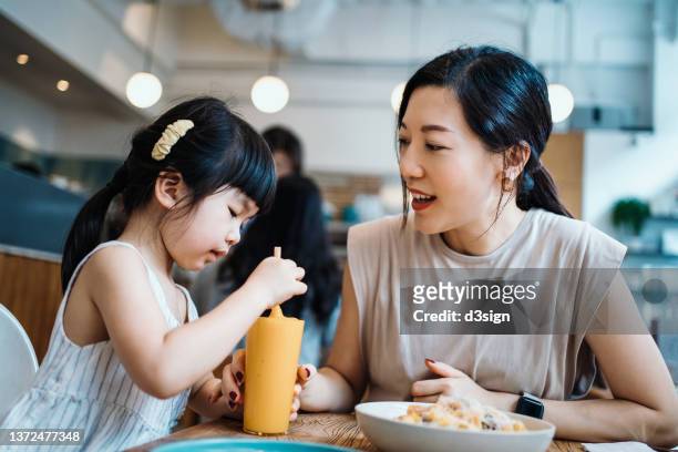 Young Asian mother and lovely little daughter having lunch together in a cafe, sharing mango smoothie and pasta. They are talking and looking at each other smiling joyfully. Mother and daughter relationship. Family and eating out lifestyle