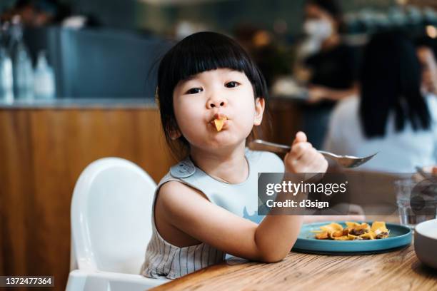 portrait of lovely little asian girl sitting on a high chair having pasta for lunch in cafe. she is making a face while looking at camera sitting at dining table. people, food and lifestyle concept - pasta geniessen stock-fotos und bilder