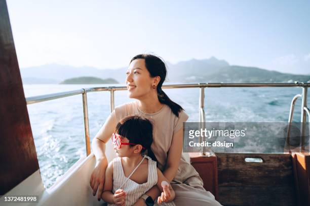 young asian mother and lovely little daughter with red sunglasses enjoying a yacht ride over nice calm sea on a summer day. looking over and relaxed at the sea. family lifestyle. enjoying outdoor fun, boat trip and sea vacations - family travel stockfoto's en -beelden