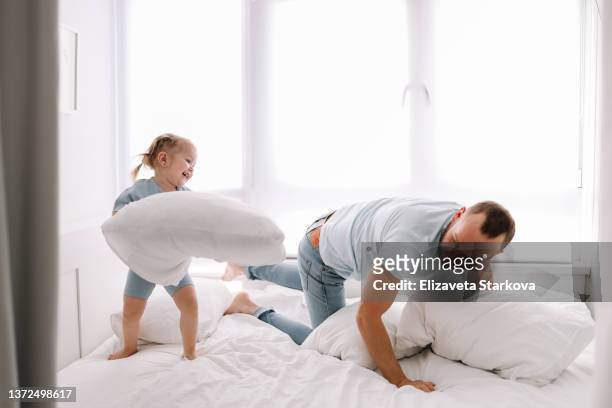a happy father and daughter have fun playing and fighting with pillows on the bed in a cozy bedroom on a weekend at home. one parent will spend time with a small child. positive masculinity - father and child and pillow fight stock pictures, royalty-free photos & images