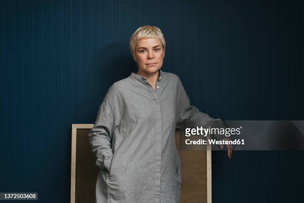 woman with short blond hair standing in front of blue wall - kunsthändler stock-fotos und bilder
