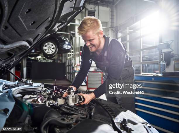 smiling young blond mechanic repairing car engine in auto shop - autowerkstatt gegenlicht stock-fotos und bilder