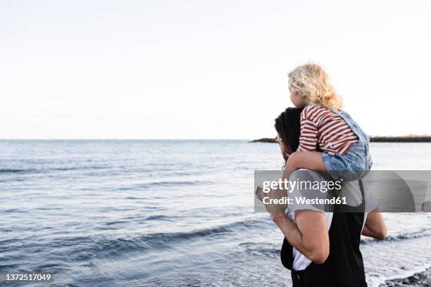 man carrying son on shoulders by seashore at beach - father son admiration stock pictures, royalty-free photos & images