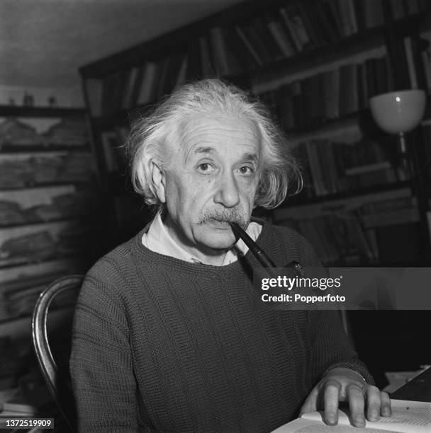 German born American theoretical physicist Albert Einstein seated smoking a pipe at a desk in his study at home on 112 Mercer Street in Princeton,...
