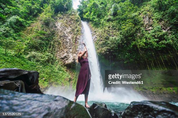 young woman standing on rock with arms raised in front of la fortuna waterfall, costa rica - costa rica women stockfoto's en -beelden