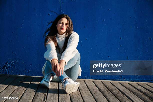 young woman sitting in front of blue wall - legs crossed at ankle stock-fotos und bilder