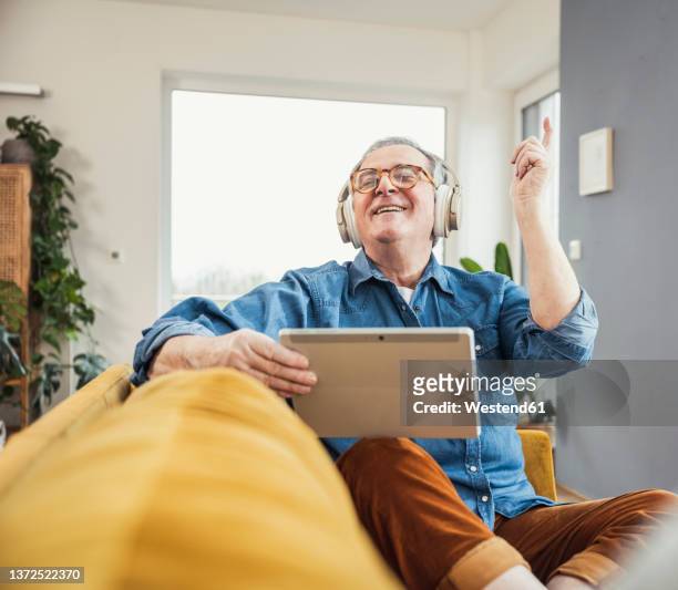 happy senior man holding tablet pc enjoying music through wireless headphones in living room - music room stockfoto's en -beelden