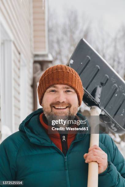 happy man wearing knit hat carrying snow shovel on shoulder in winter - snow shovel man stock pictures, royalty-free photos & images