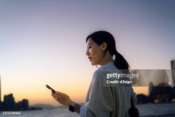 young asian businesswoman standing at harbour, managing banking and finance with mobile app on smartphone on the go. with the silhouette of urban cityscape over sunset sky in the background. smart banking with technology - forex fotografías e imágenes de stock