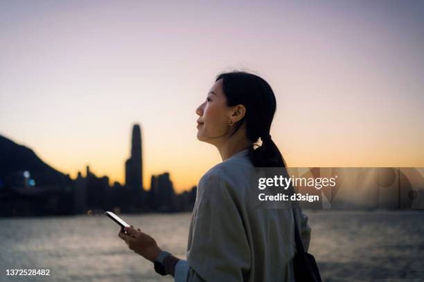 young asian businesswoman standing at harbour, looking away while using smartphone. with the silhouette of urban cityscape over sunset sky in the background. business on the go - will call stock pictures, royalty-free photos & images