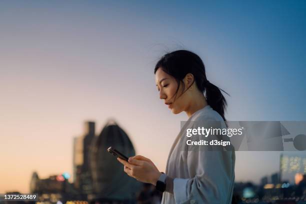 modern young asian woman walking in the city text messaging on smartphone on the go, against urban city scene with corporate skyscrapers at sunset. lifestyle and technology - go red for women fotografías e imágenes de stock