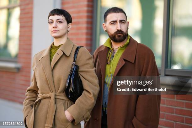 Guest wears silver and gold earrings, a green embossed / cut-out flower print pattern high neck t-shirt, a beige long belted coat, a black shiny...