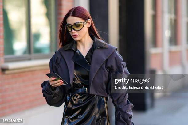 Guest wears yellow and black sunglasses, a black shiny varnished leather long coat, a dark cropped oversized jacket, outside the Fendi fashion show,...