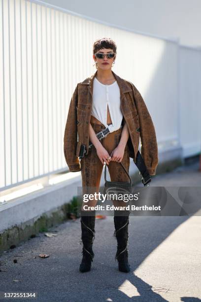 Guest wears beige and brown sunglasses, silver earrings, a white ribbed buttoned cropped t-shirt, a brown leather oversized jacket, a black faded...