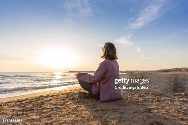 silhouette of mid adult woman practicing yoga lotus position at beach. - sunrise yoga stock pictures, royalty-free photos & images