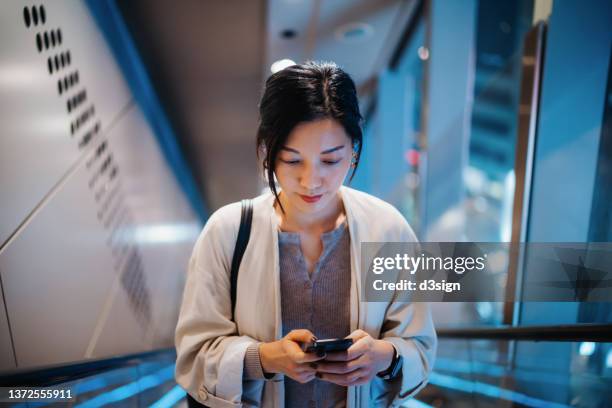 confident young asian businesswoman reading emails from business clients on smartphone while riding on escalator in modern office building in the evening. business lifestyle with technology. business on the go - airport asian worker stock-fotos und bilder