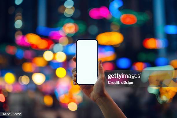 cropped shot of woman's hand holding up a smartphone against illuminated and multi-coloured bokeh lights background in the city at night - multi devices stockfoto's en -beelden