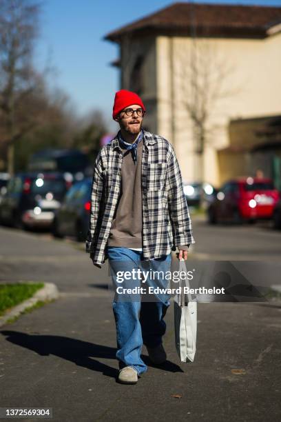 Guest wears a red wool beanie, black glasses, a navy blue and white print pattern scarf, a brown sweater, a beige and black checkered print pattern...