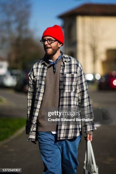 Guest wears a red wool beanie, black glasses, a navy blue and white print pattern scarf, a brown sweater, a beige and black checkered print pattern...