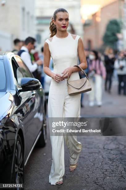Guest wears gold earrings, a white latte with transparent shoulder sleeveless flared jumpsuit, silver and diamonds rings, a beige shiny leather with...