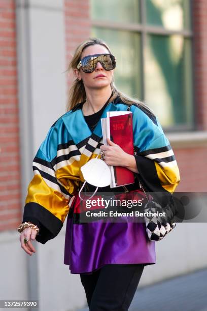 Guest wears brown ski sunglasses, a black t-shirt, a blue / beige / black / gold / red / purple shiny silk pattern belted jacket, a black and white...