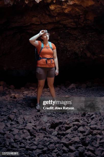 a young woman stands at the bottom of a cavernous lava tube and looks up towards a shaft of light - pothole stock pictures, royalty-free photos & images