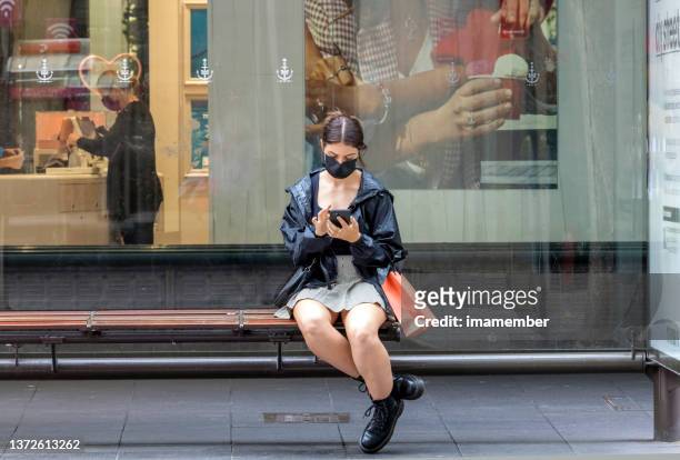 junge frau mit geschützter gesichtsmaske sitzt in der bushaltestelle und benutzt ihr smartphone, hintergrund mit kopie sapce - the face of australia photo call stock-fotos und bilder