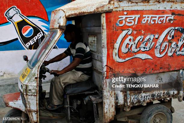 An Indian beverage delivery man, driving a dilapidated vehicle adorned with a hand-painted Coca-Cola logo, drives past a mural of a Pepsi bottle in...