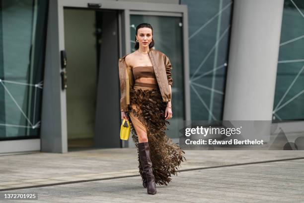 Mary Leest wears gold earrings, a gold chain necklace, a brown shoulder-off / cropped top from Max Mara, a dark brown shiny leather bomber coat from...