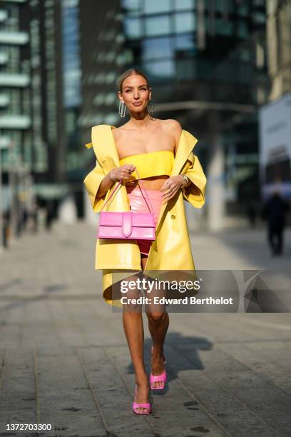 Leonie Hanne wears large gold square pendant earrings, a yellow shoulder-off / cropped top, a yellow shiny leather oversized coat, a high waist pink...