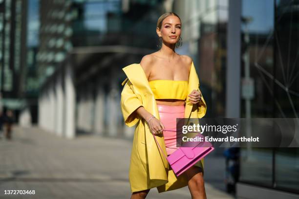 Leonie Hanne wears large gold square pendant earrings, a yellow shoulder-off / cropped top, a yellow shiny leather oversized coat, a high waist pink...