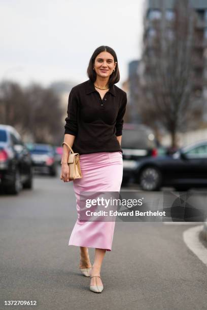 Kate Darma wears gold earrings, a gold large necklace, a black polo shirt from Prada, a beige shiny leather handbag, a pale pink satin silk midi tube...