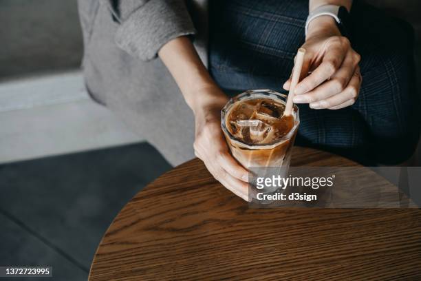 high angle, cropped shot of young asian woman holding a glass of iced coffee with eco friendly straw on the wooden table enjoying a coffee break in coffee shop - iced coffee foto e immagini stock