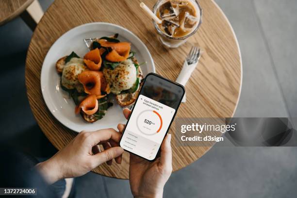 overhead view of young asian woman using fitness plan mobile app on smartphone to tailor make her daily diet meal plan, checking the nutrition facts and calories intake of her meal, eggs benedict with smoked salmon in cafe. maintaining a healthy diet - meal plan stock-fotos und bilder