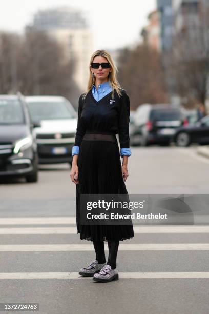 Natalia Verza wears black sunglasses, a black and silver earring from Prada, a blue shirt, a black V-neck wool pullover from Prada, a black...