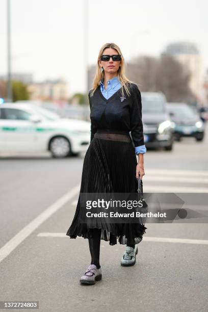 Natalia Verza wears black sunglasses, a black and silver earring from Prada, a blue shirt, a black V-neck wool pullover from Prada, a black...