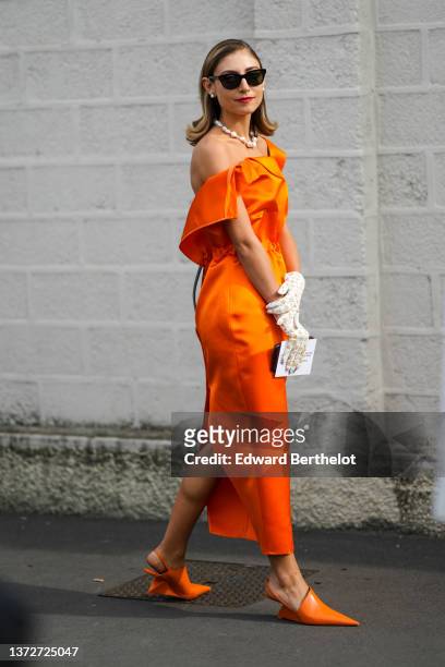 Jenny Walton wears black sunglasses, pearl earrings, pearls necklace, an orange shiny shoulder-off / ruffled / belted slit midi dress, a white with...